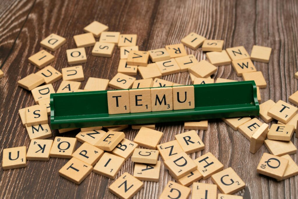 Wooden Scrabble tiles spelling TEMU on a green rack with scattered tiles on a wooden surface.